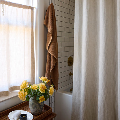 Yellow roses in a ceramic vase on a wicker tray, beige sheer curtains, brown towel, white subway tiles, brass tub fixtures.
