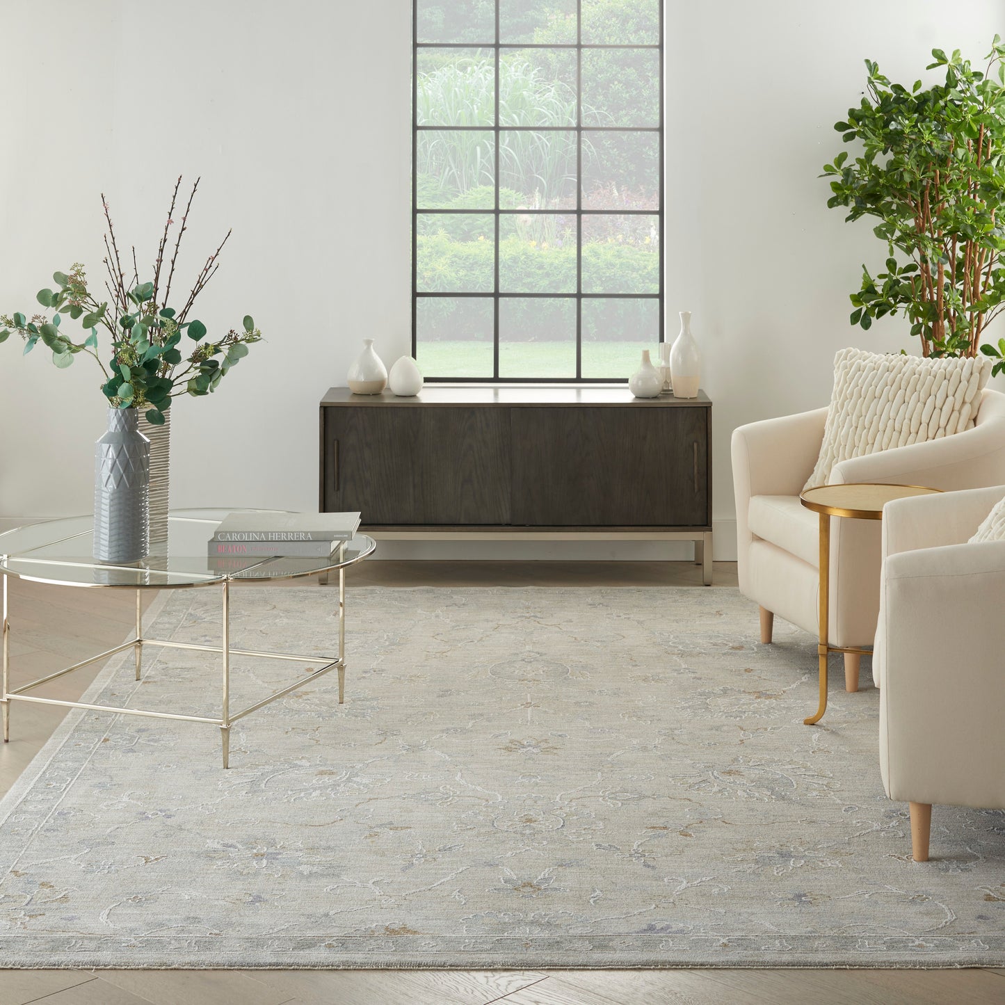 Bright modern living room with beige armchairs, glass coffee table, grey rug, dark wood console, grid window, leafy plants.
