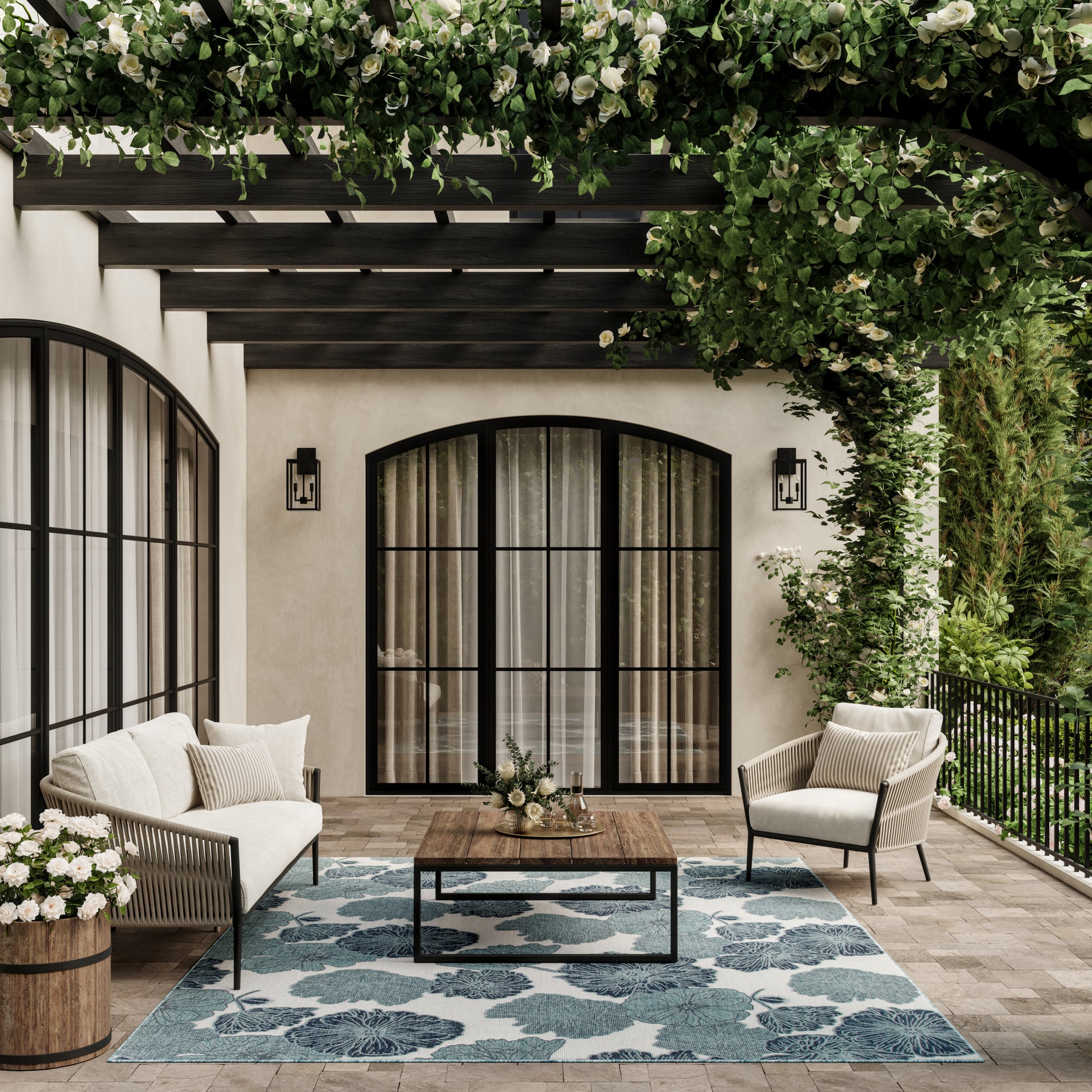 Outdoor patio with cream cushioned seating, wooden coffee table, blue floral rug, arched glass doors, pergola and lush greenery.