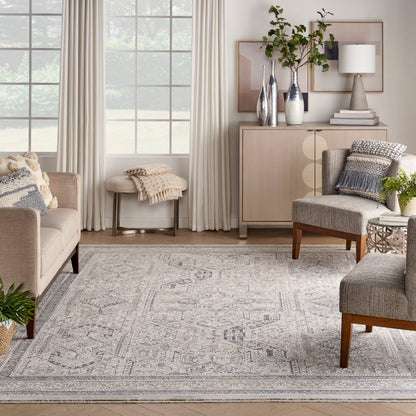 Neutral living room with beige sofas, gray geometric rug, wood console, vases, plants, and soft natural light from windows.