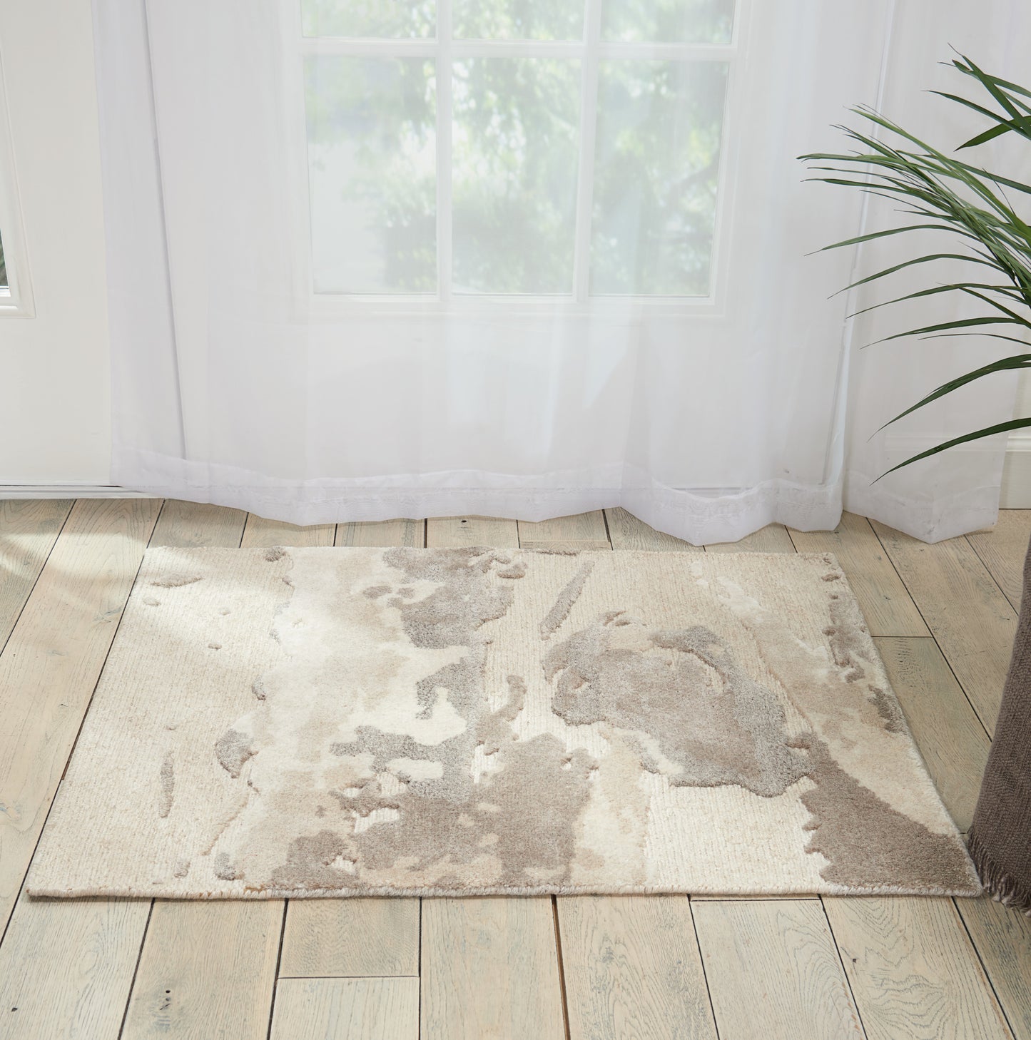 Beige abstract area rug on light wood floor beside sheer white curtains and a green potted plant.