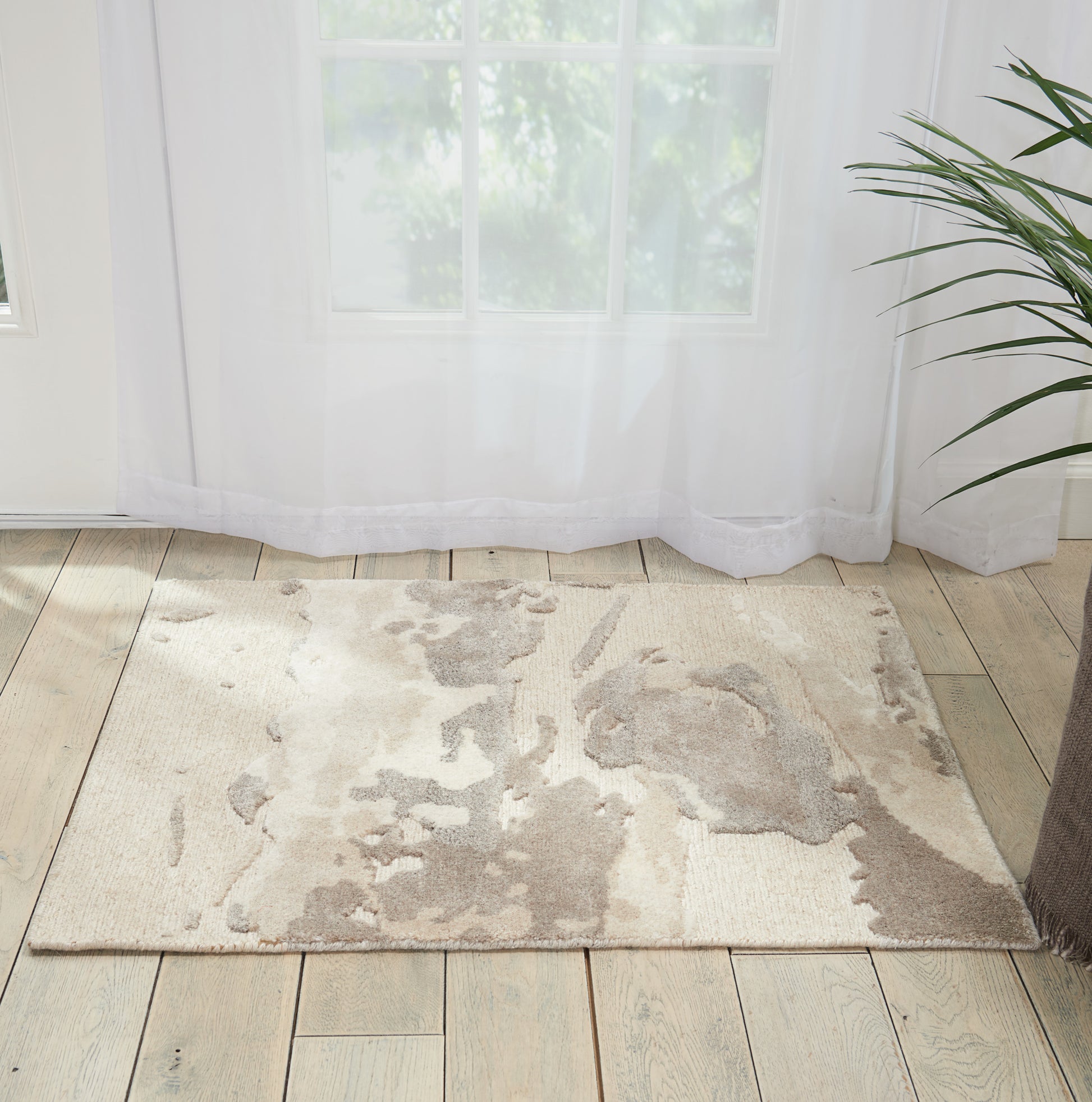 Beige abstract area rug on light wood floor beside sheer white curtains and a green potted plant.
