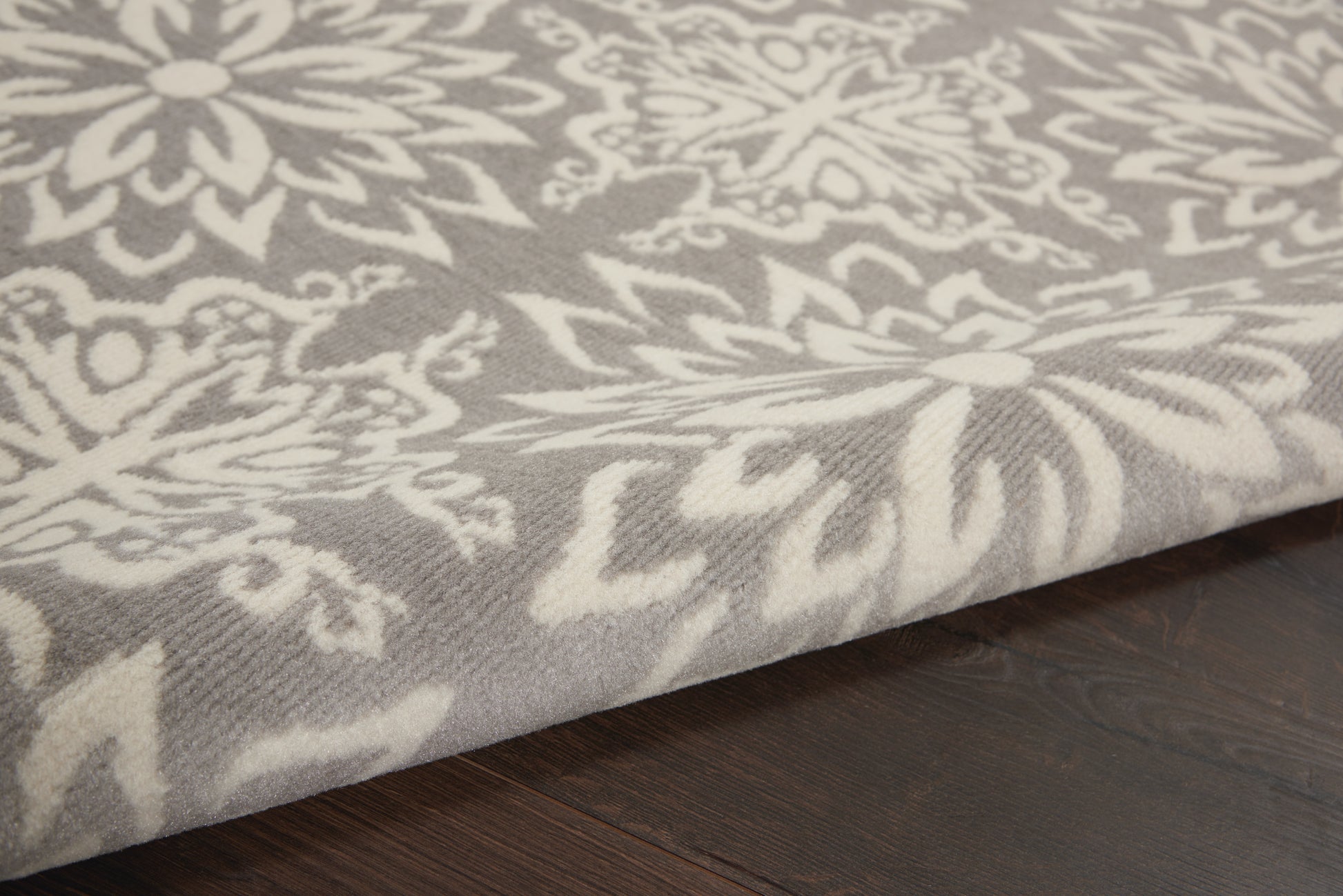 Close-up of a gray-white damask-pattern rug on dark wood floor with ornate floral motifs.