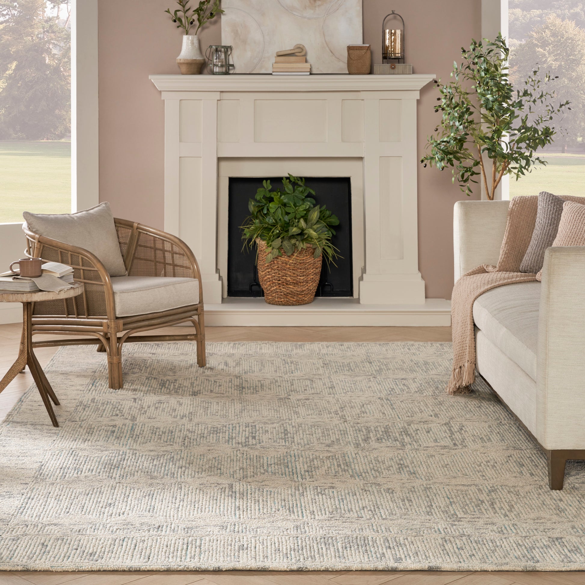 Cozy neutral living room with white mantel, beige sofa, rattan chair, patterned area rug, potted plant by fireplace.