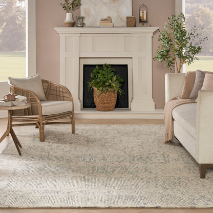 Cozy neutral living room with white mantel, beige sofa, rattan chair, patterned area rug, potted plant by fireplace.