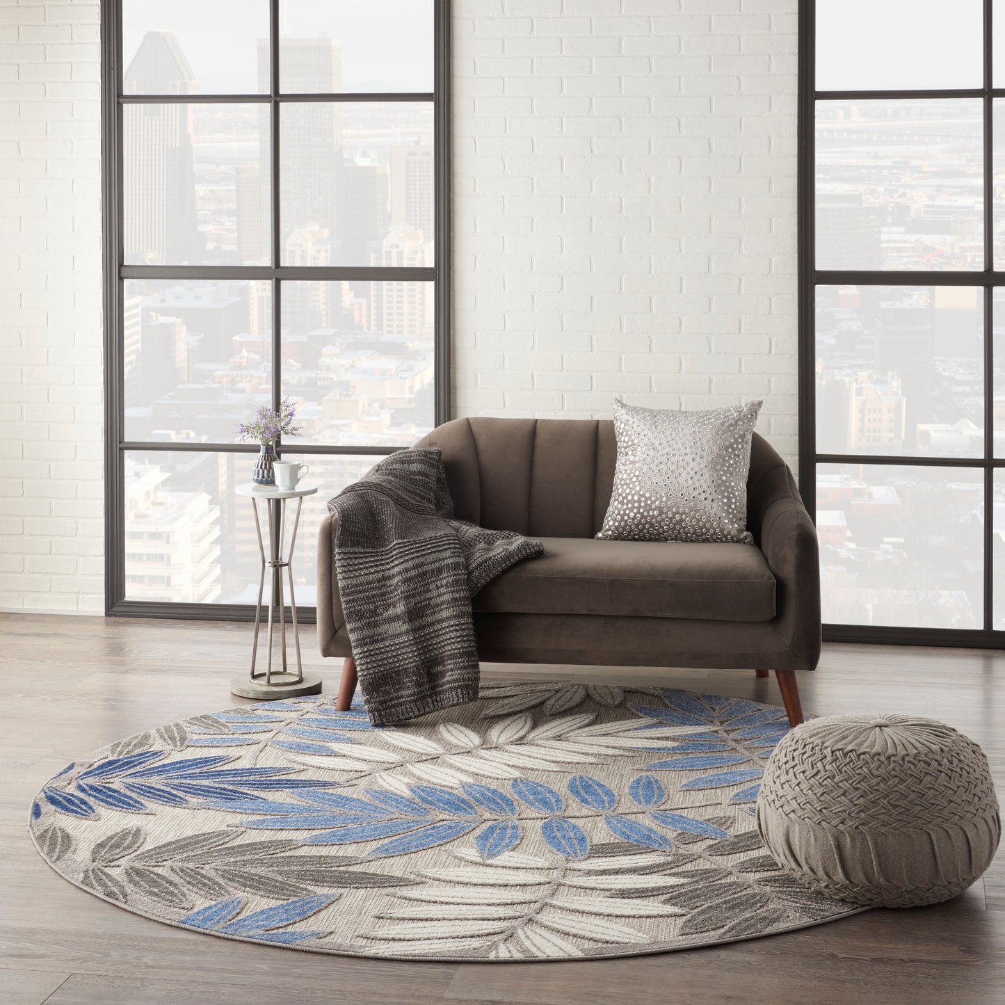 Brown mid-century sofa with grey throw and metallic pillow on a round blue-gray leaf rug, knit pouf and city-view windows.