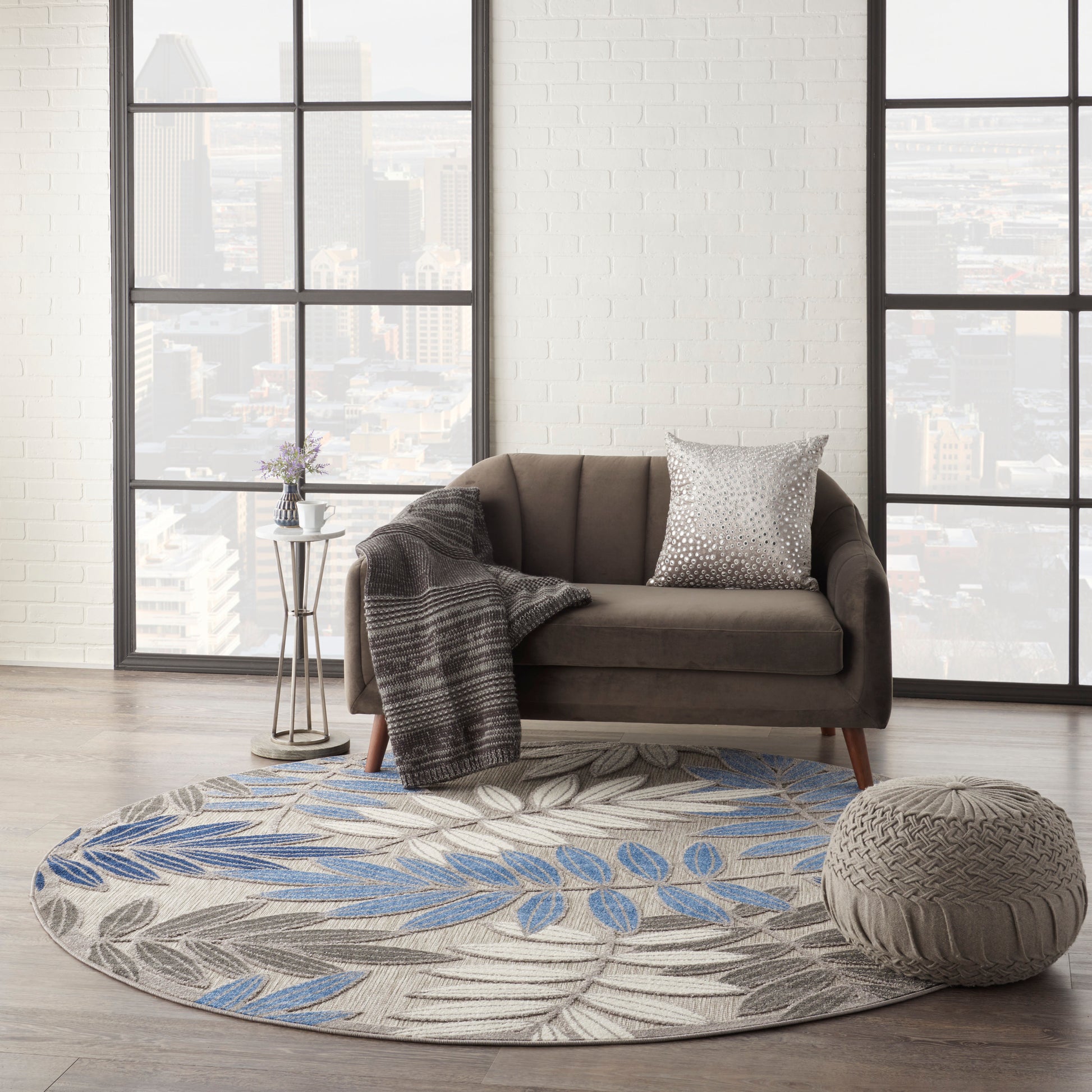 Brown mid-century sofa with grey throw and metallic pillow on a round blue-gray leaf rug, knit pouf and city-view windows.