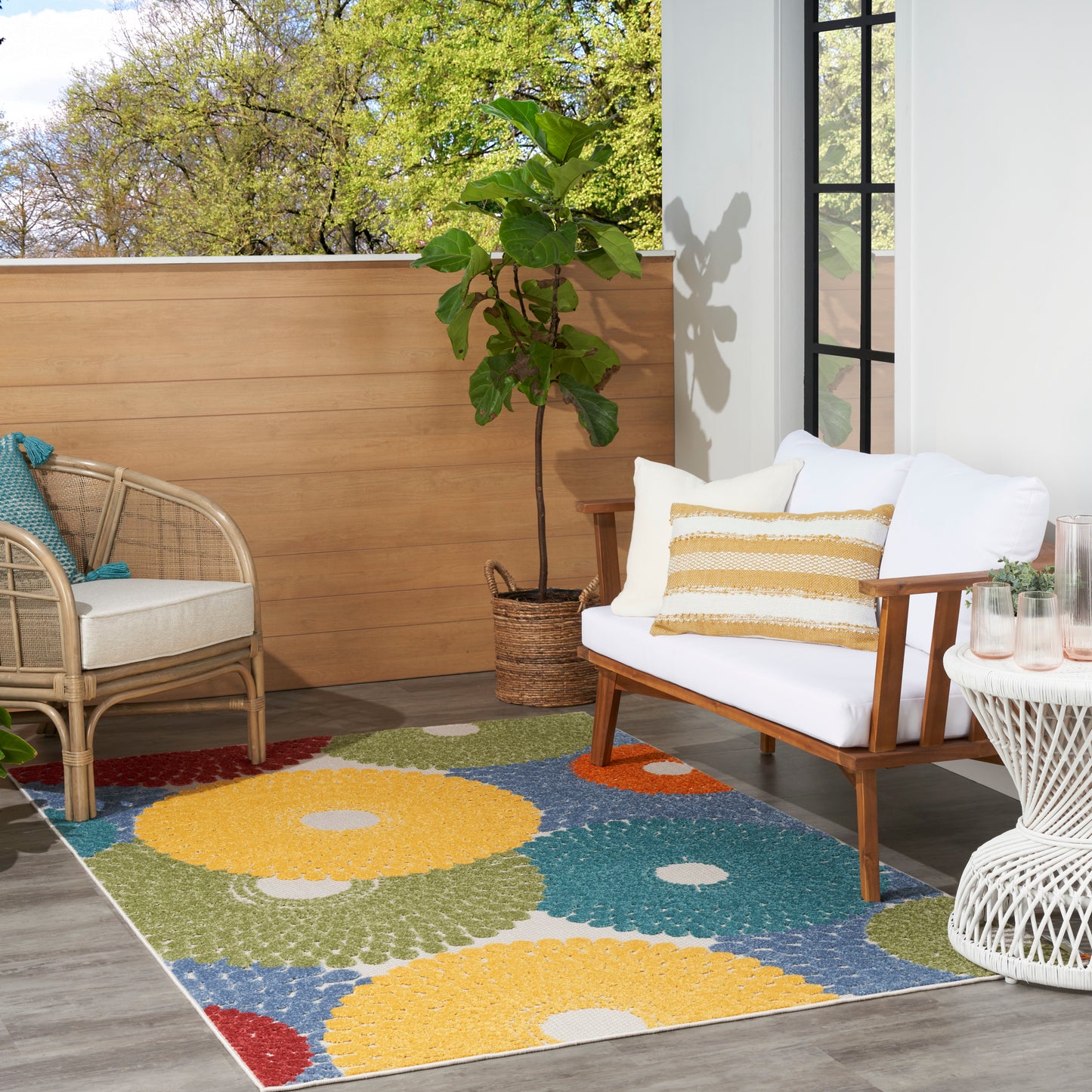 Cozy balcony with colorful rectangular rug, white cushioned loveseat, rattan chair, potted plant and wicker side table.