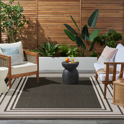 Cozy outdoor seating with wood-framed chairs, beige cushions, black round side table, lush planters, and a geometric rug.