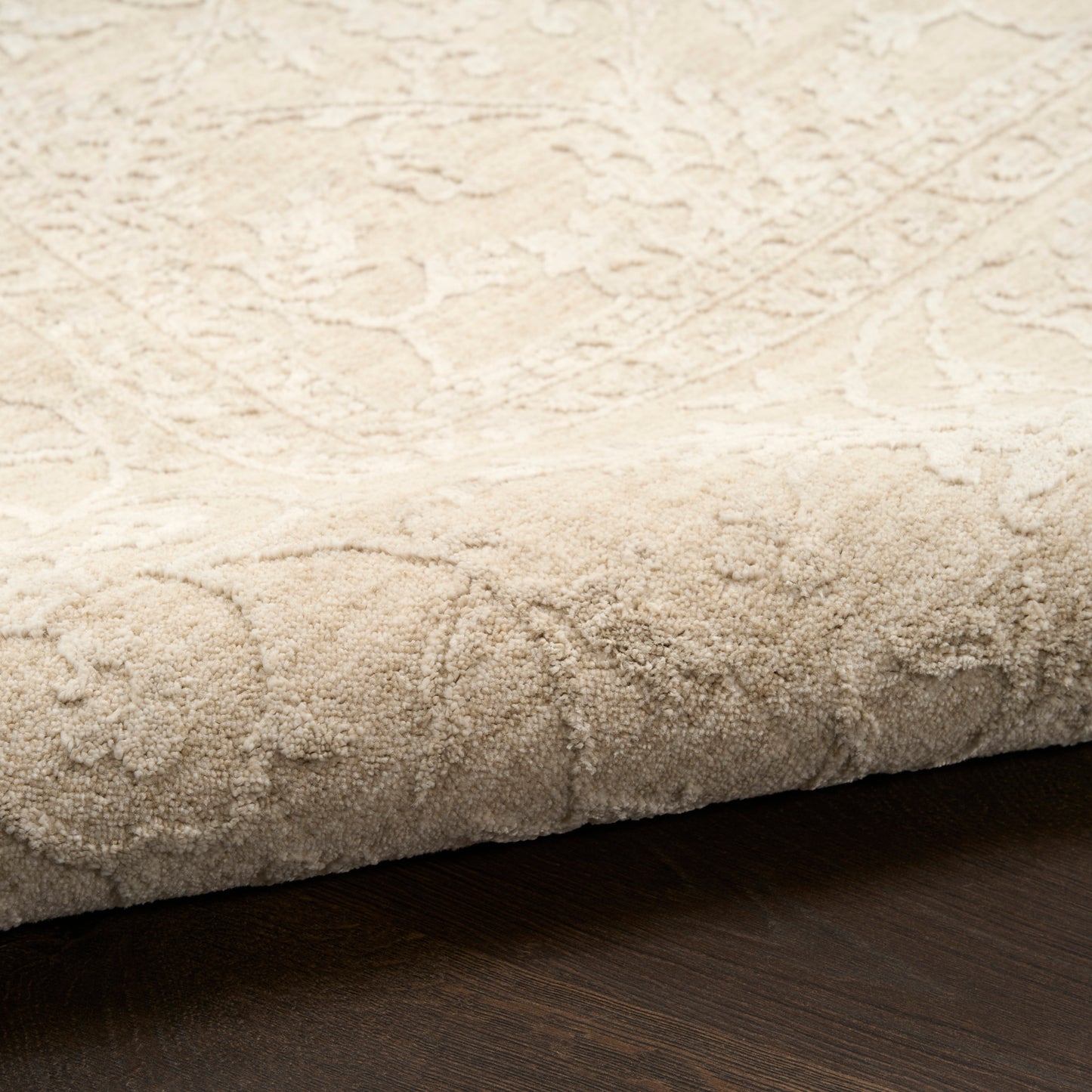 Cream textured area rug with subtle damask pattern and plush pile, edge folded over on dark wood floor.
