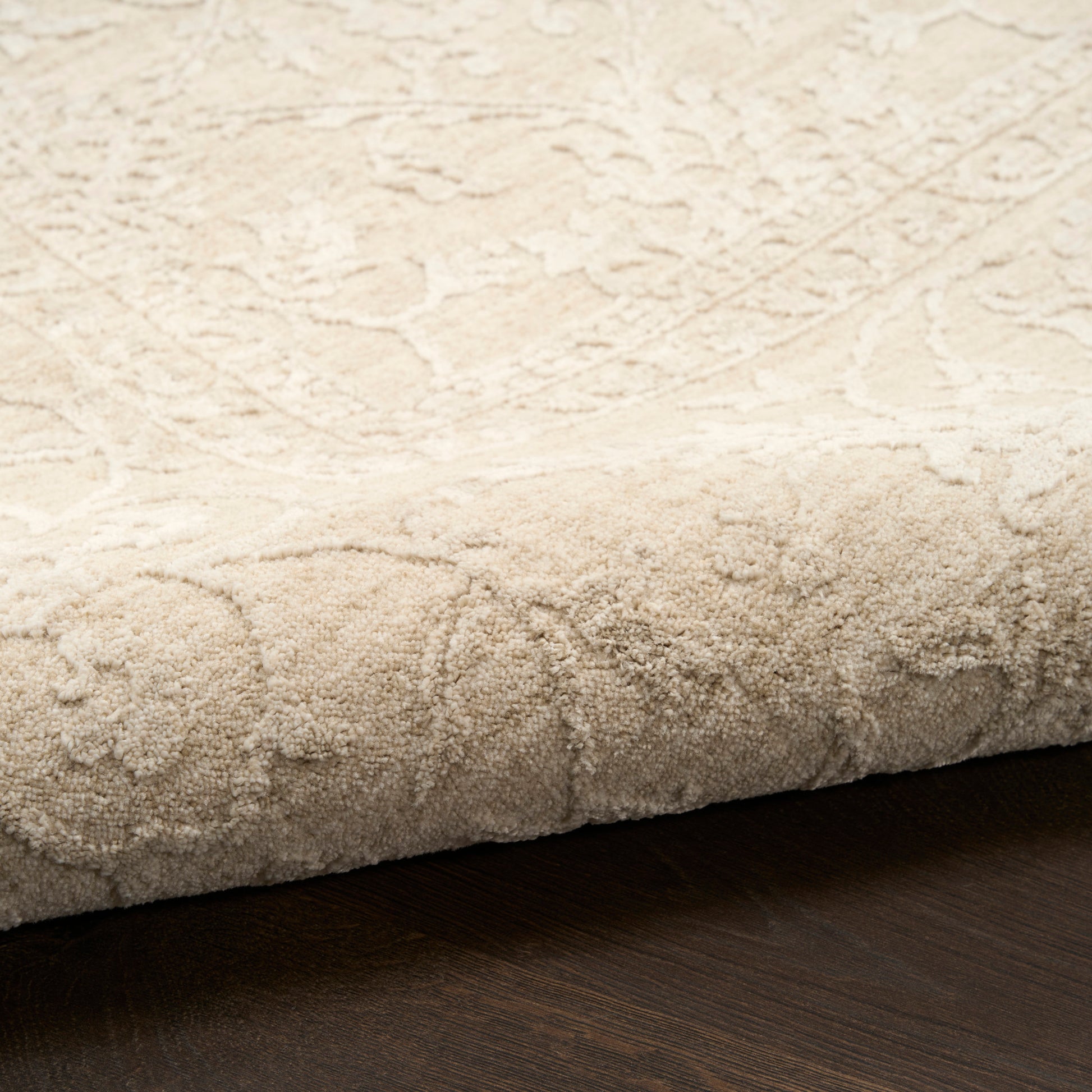 Cream textured area rug with subtle damask pattern and plush pile, edge folded over on dark wood floor.