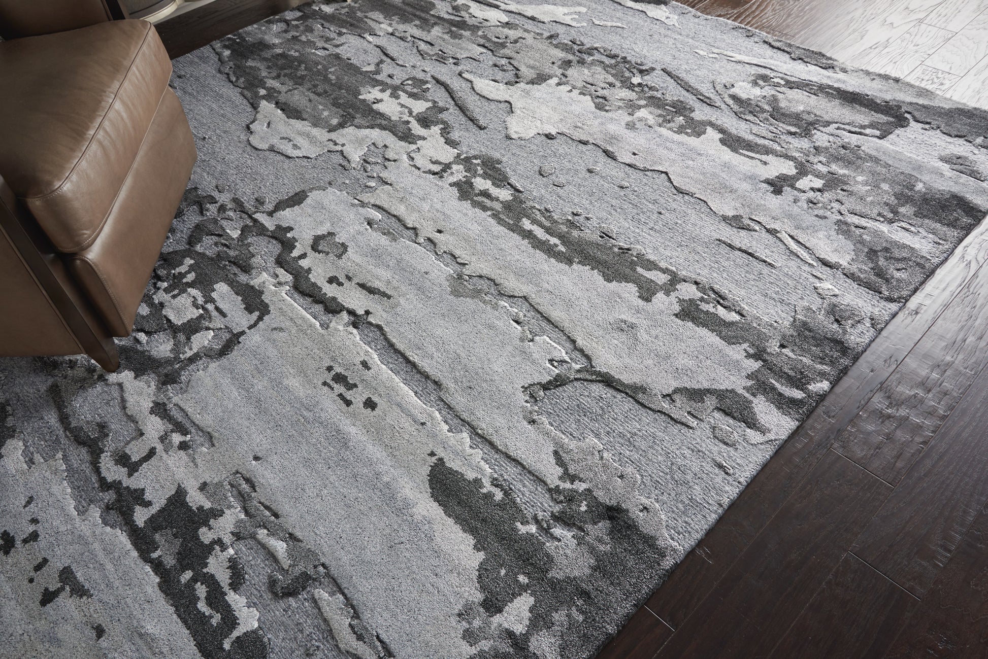Abstract gray & charcoal rug with irregular patches on dark wood floor; brown leather chair corner.