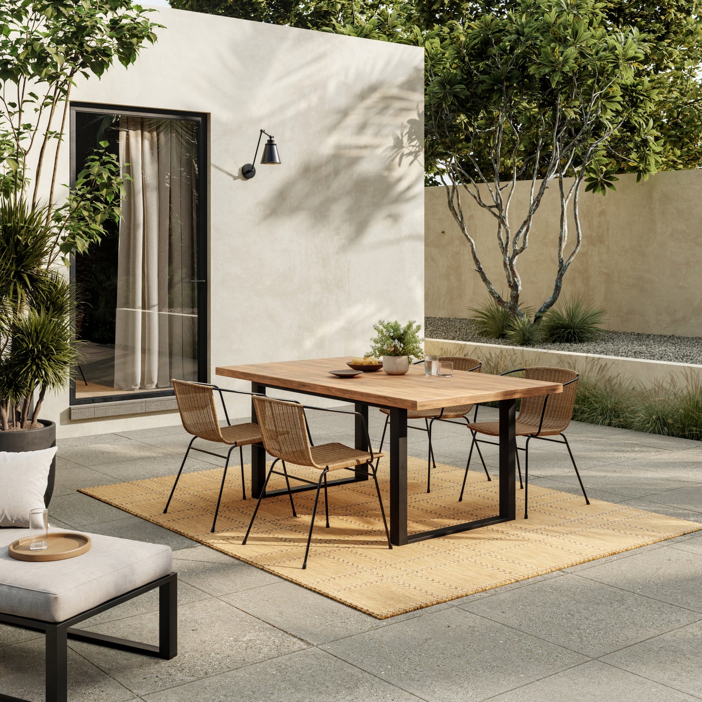 Outdoor dining set: rectangular wooden table with black metal legs and woven rattan chairs on a beige rug in a courtyard.