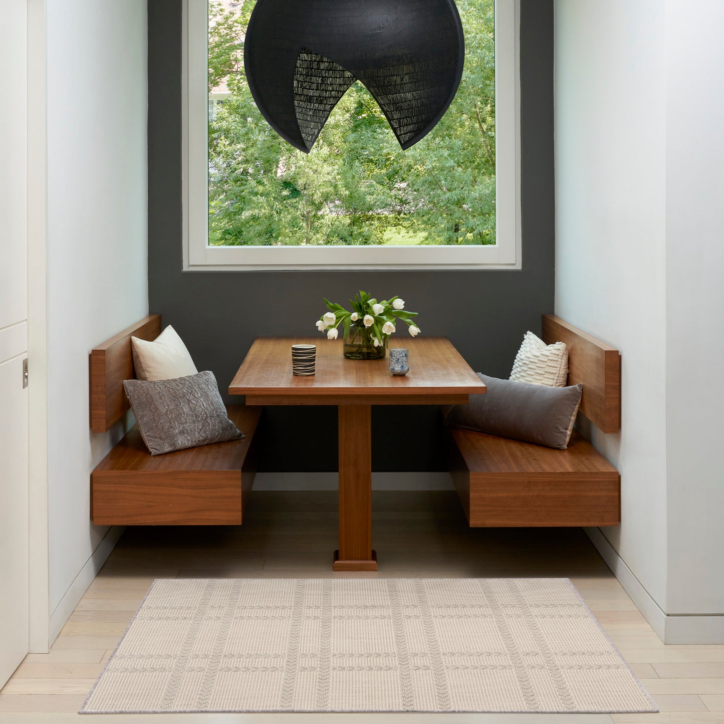 Cozy walnut dining nook with corner benches, square pedestal table, black curved pendant lamp, and a vase of flowers.