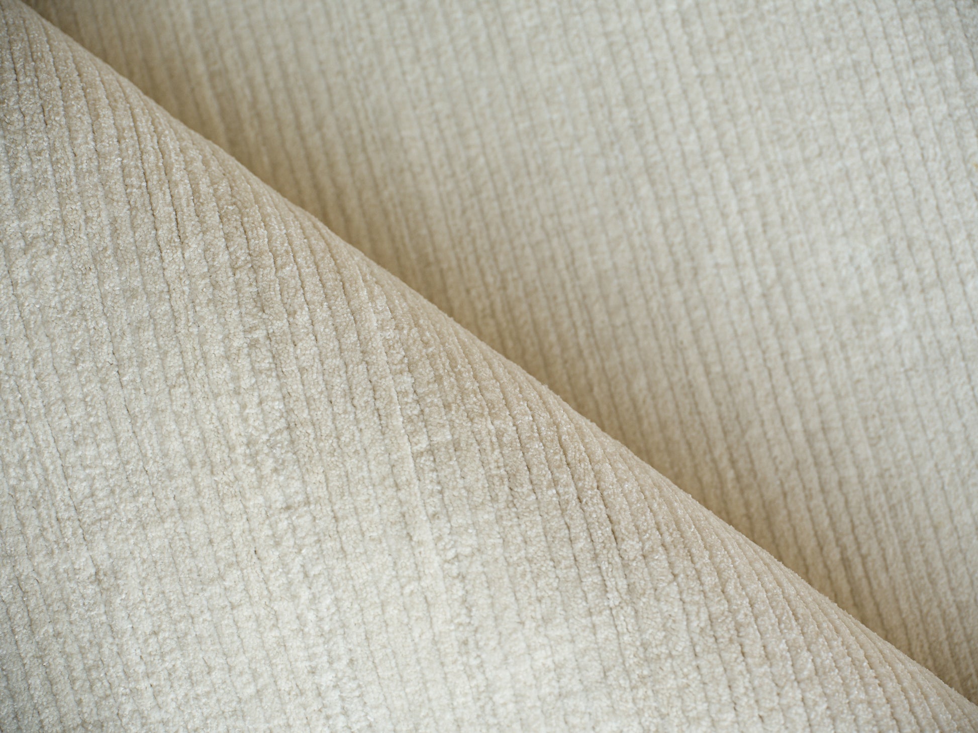 Beige corduroy fabric close-up with soft ridges and fold lines; ideal for upholstery or apparel.