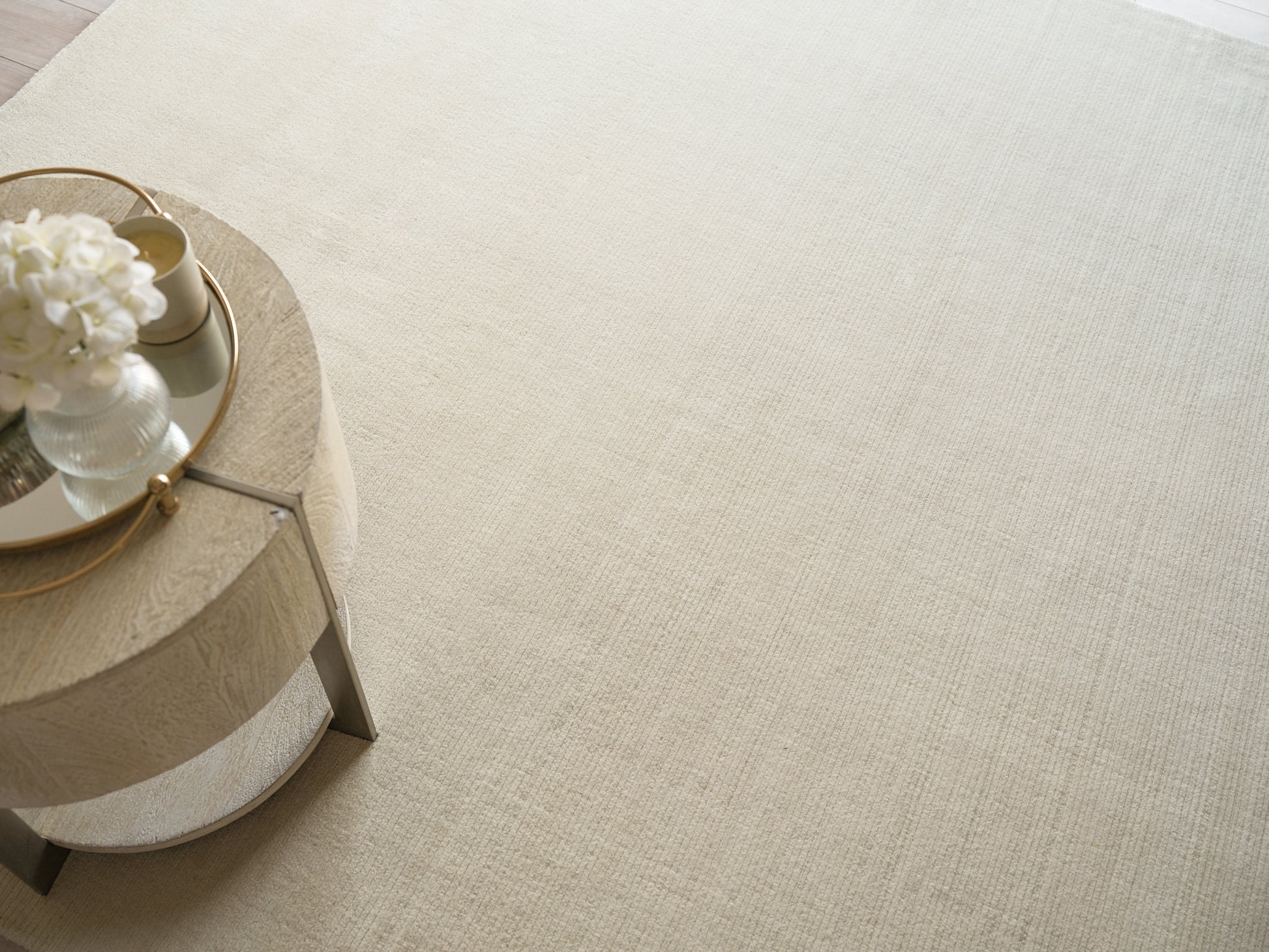 Round light-wood coffee table with metal legs, gold-rimmed mirrored tray and white bouquet on a beige rug.