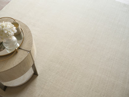 Round light-wood coffee table with metal legs, gold-rimmed mirrored tray and white bouquet on a beige rug.