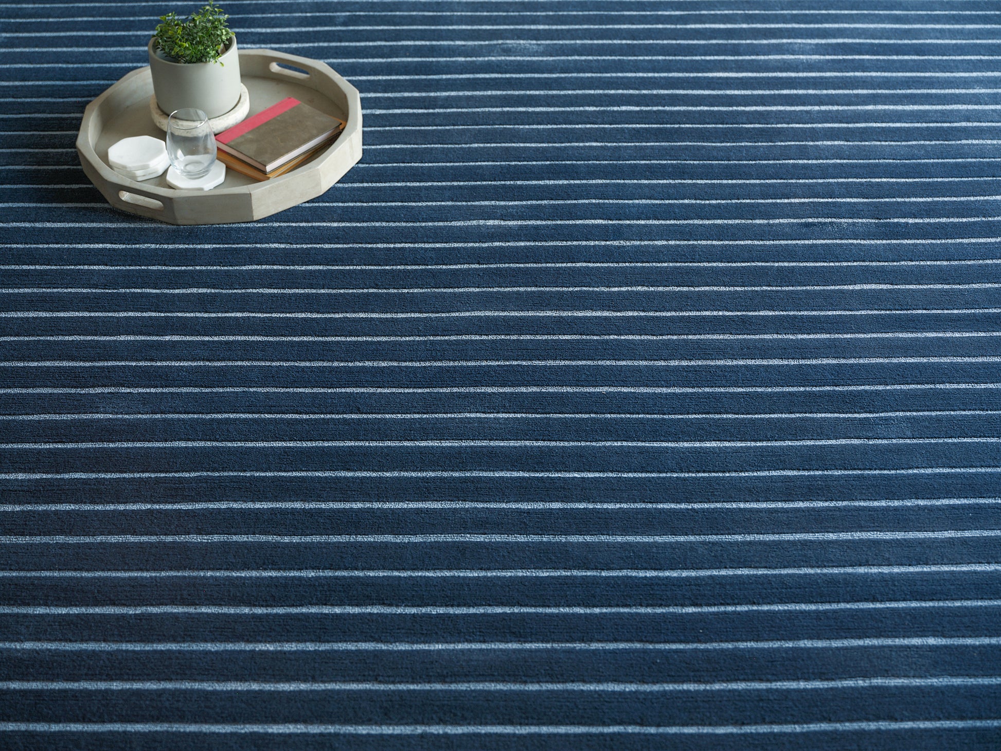 Blue striped rug with a circular beige wooden tray holding a potted plant, a glass and books.
