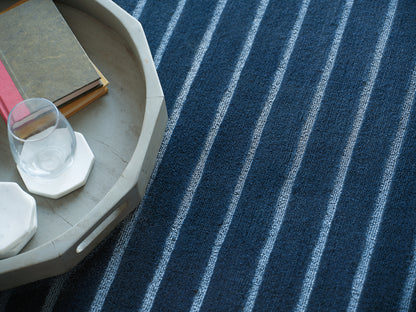 Gray oval tray with a stack of books and a glass on a white hex coaster, set on a blue striped rug.