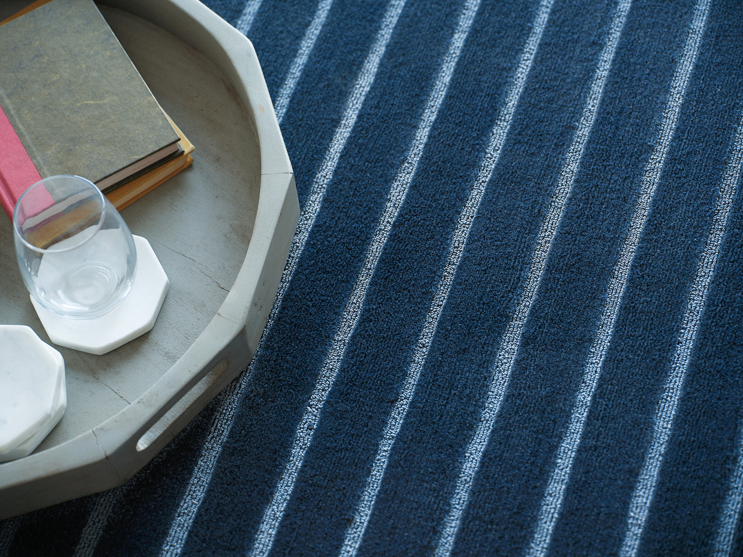 Round gray wooden tray with glass of water on white hex coaster, stacked books, on blue striped rug.