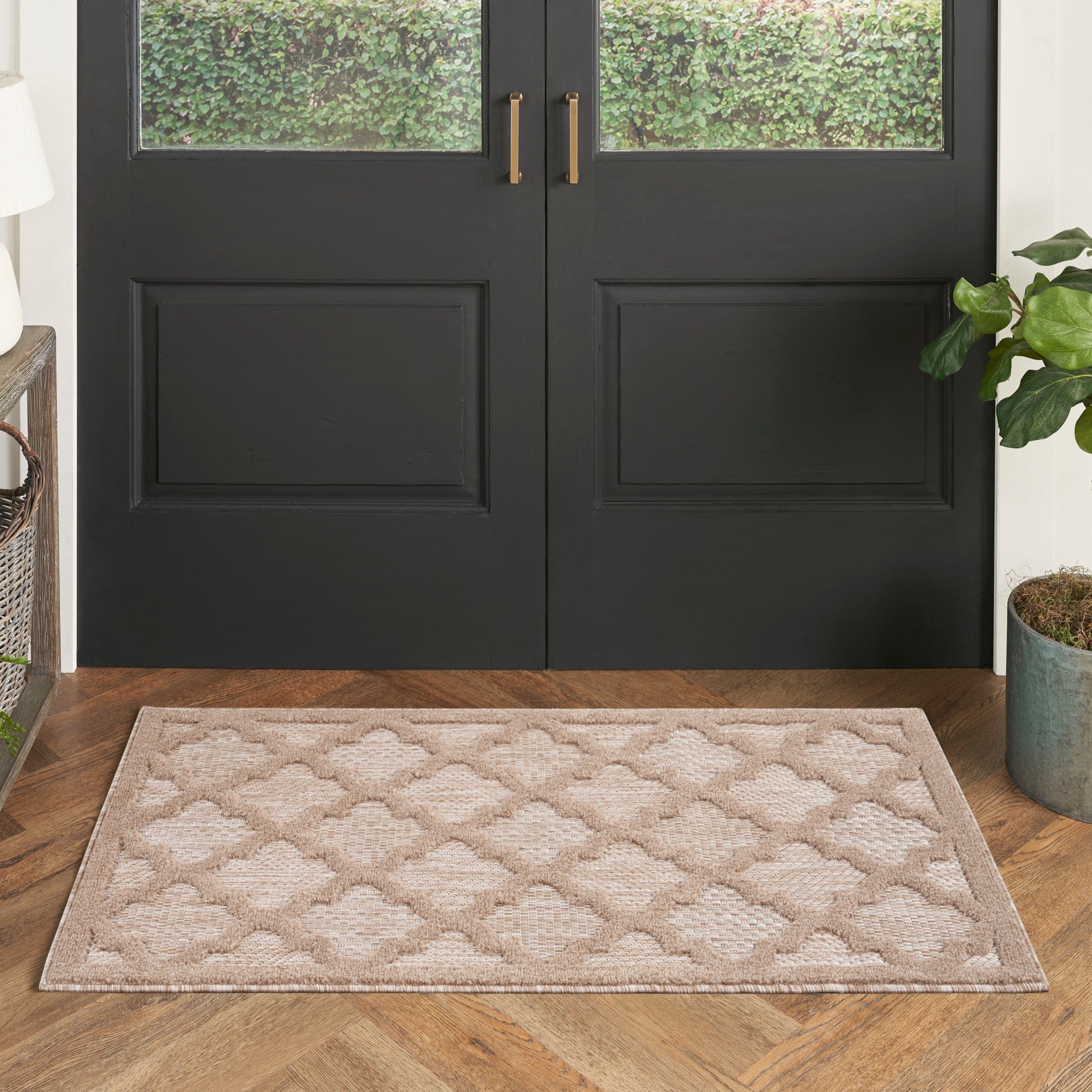 Beige entryway mat with trellis pattern on a wooden floor in front of black double doors with gold handles.