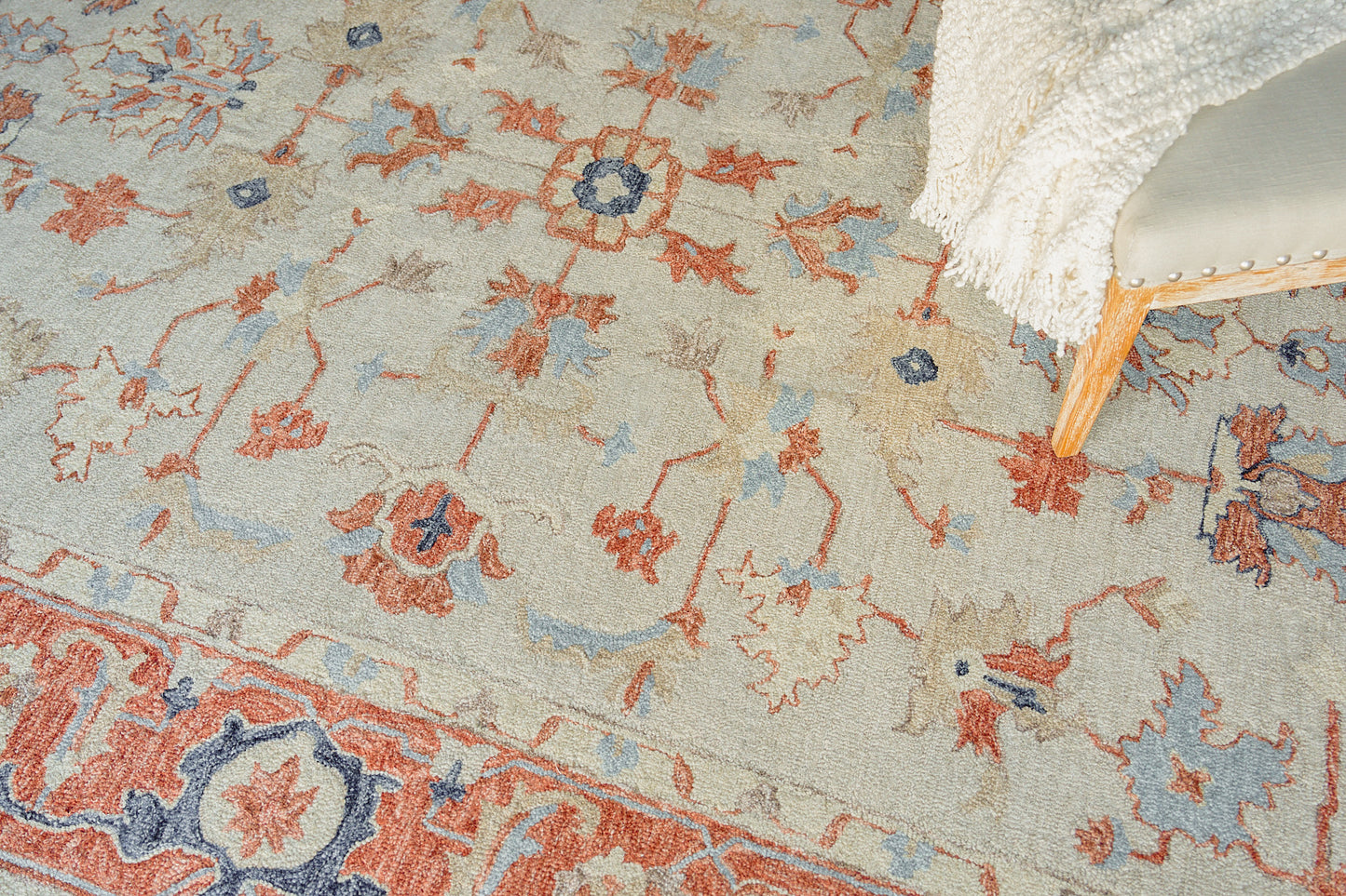 Pastel area rug with blue and peach floral motifs on beige; corner of a light wood chair with white fringe throw.