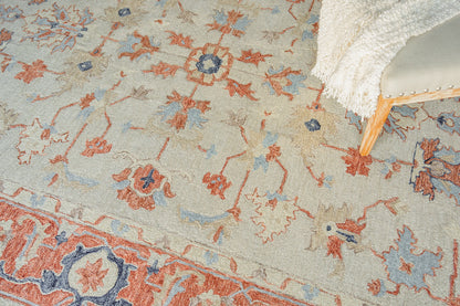 Pastel area rug with blue and peach floral motifs on beige; corner of a light wood chair with white fringe throw.