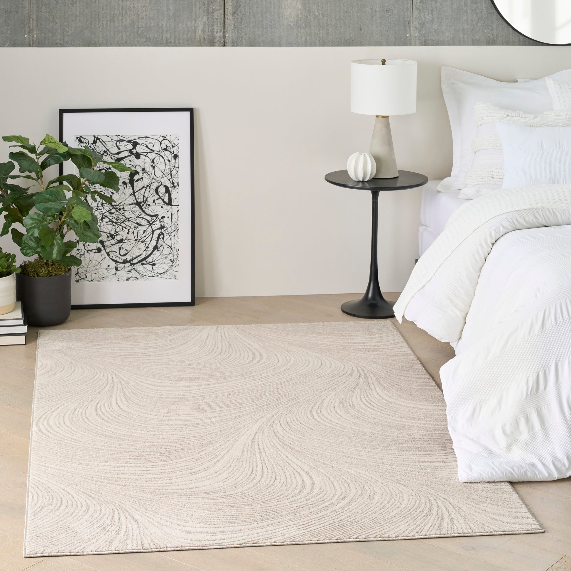 Beige swirl rug on light wood floor beside white bed; black round side table with white lamp, plant, and abstract art.