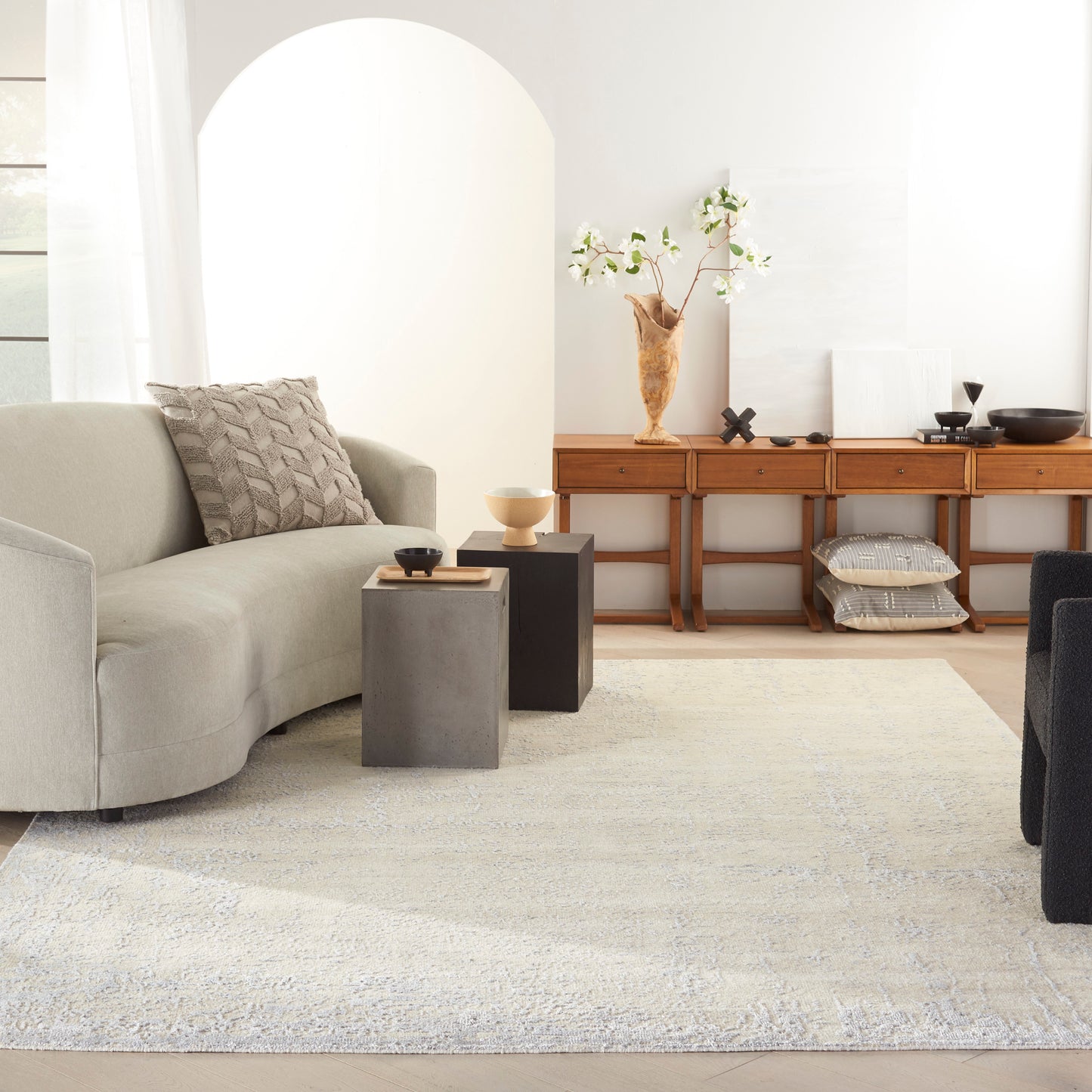 Minimalist neutral living room with beige sofa, concrete square side tables, wooden console, vase of white flowers, pale rug.
