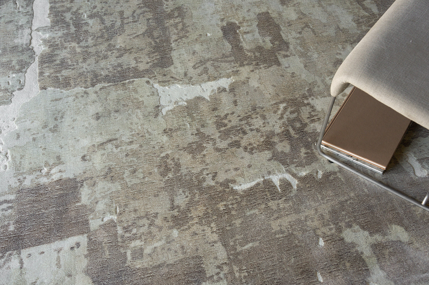 Close-up of a textured gray-beige rug with irregular patches, beside a chrome-legged chair with light beige upholstery.
