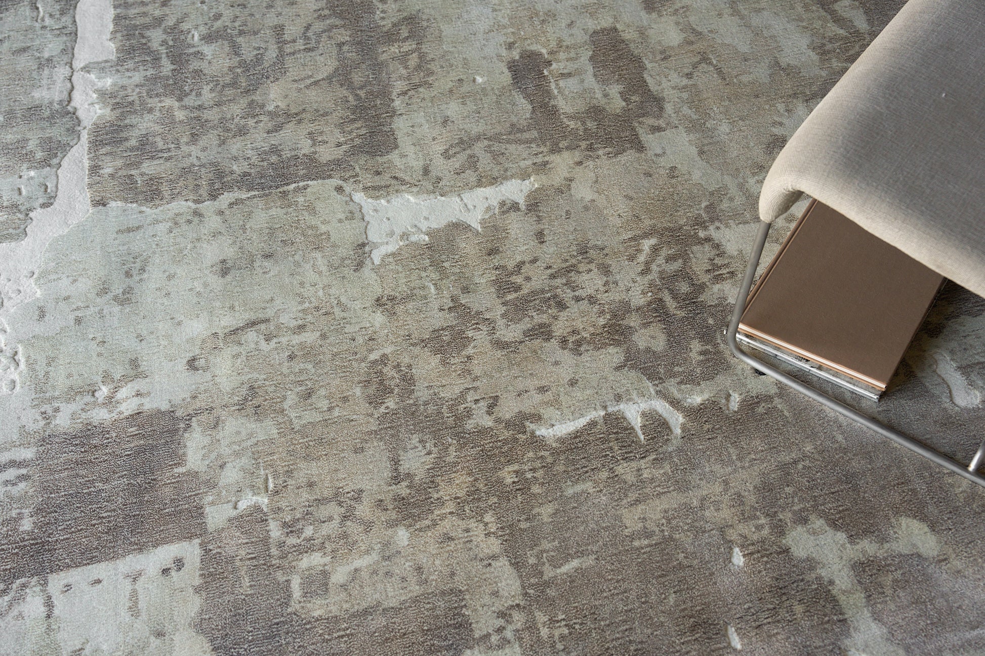 Close-up of a textured gray-beige rug with irregular patches, beside a chrome-legged chair with light beige upholstery.