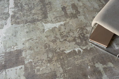 Close-up of a textured gray-beige rug with irregular patches, beside a chrome-legged chair with light beige upholstery.