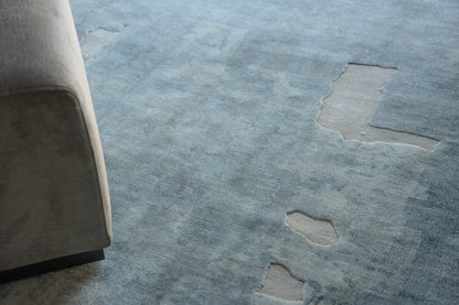 Blue distressed area rug with light beige worn patches beside a gray upholstered sofa.