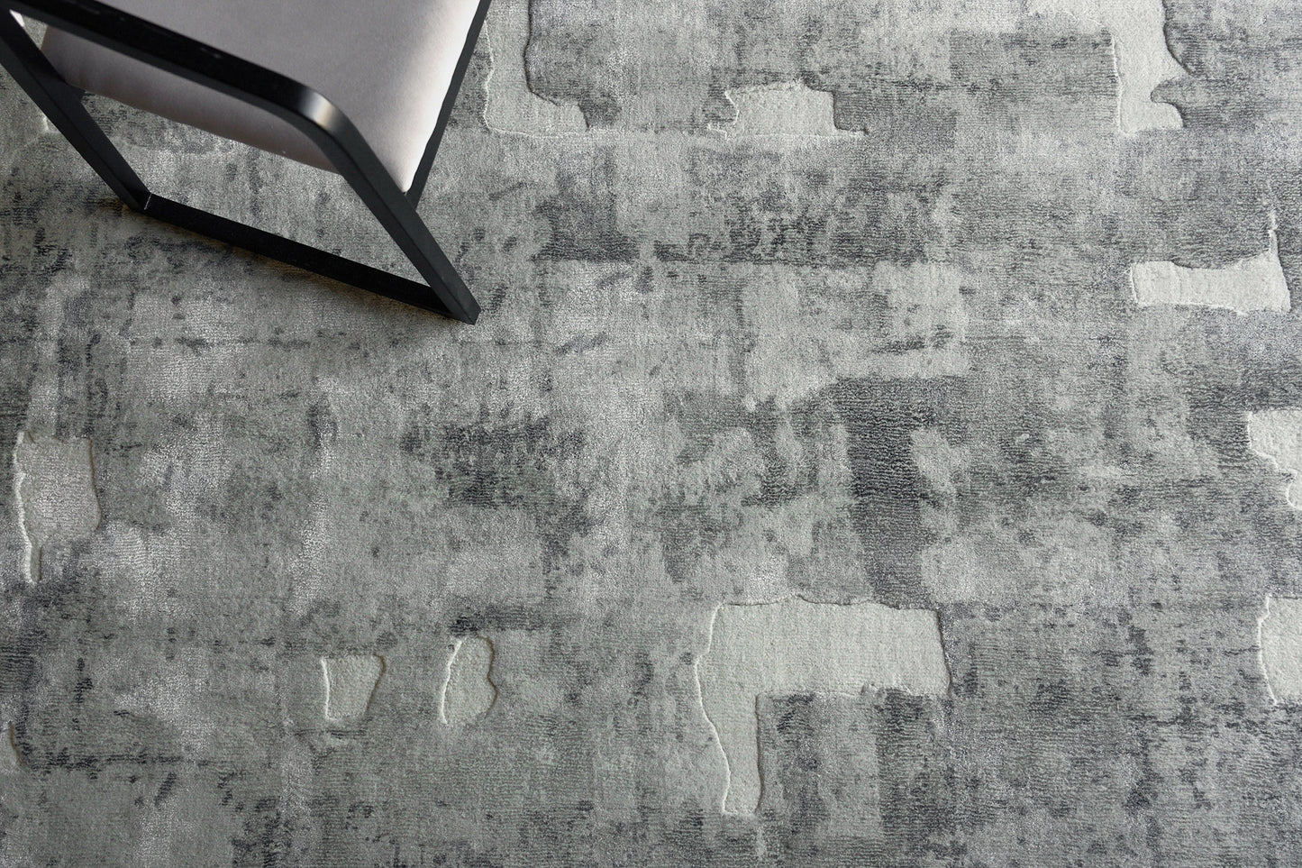 Gray textured rug with irregular lighter patches, seen with a black metal-framed chair in the corner.