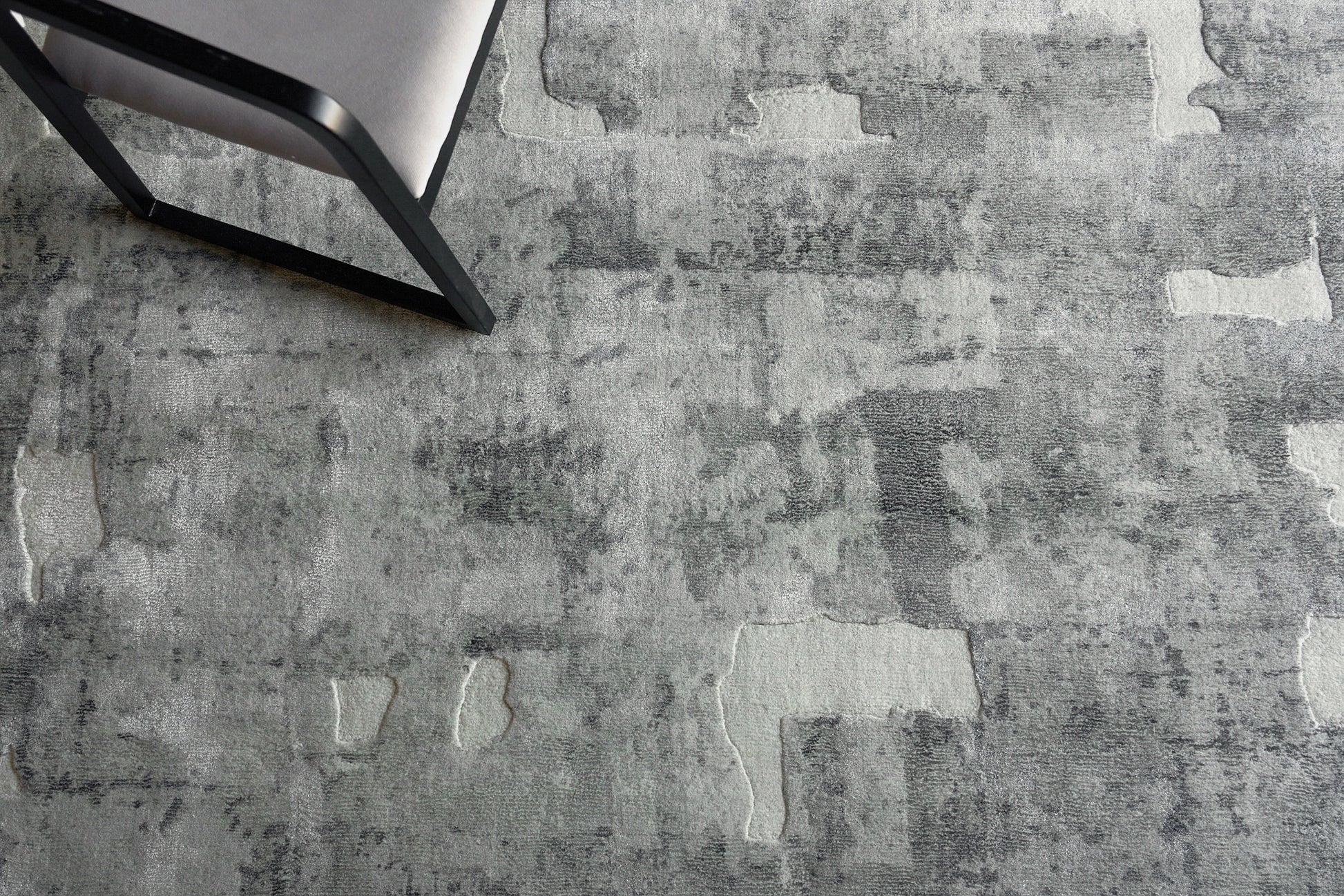 Gray textured rug with irregular lighter patches, seen with a black metal-framed chair in the corner.