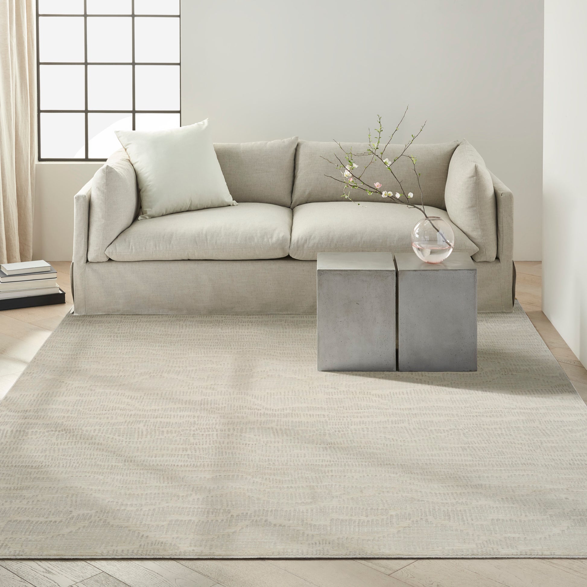 Beige fabric sofa with cream throw pillow, large textured beige rug, concrete block side tables, glass vase with blossoms.