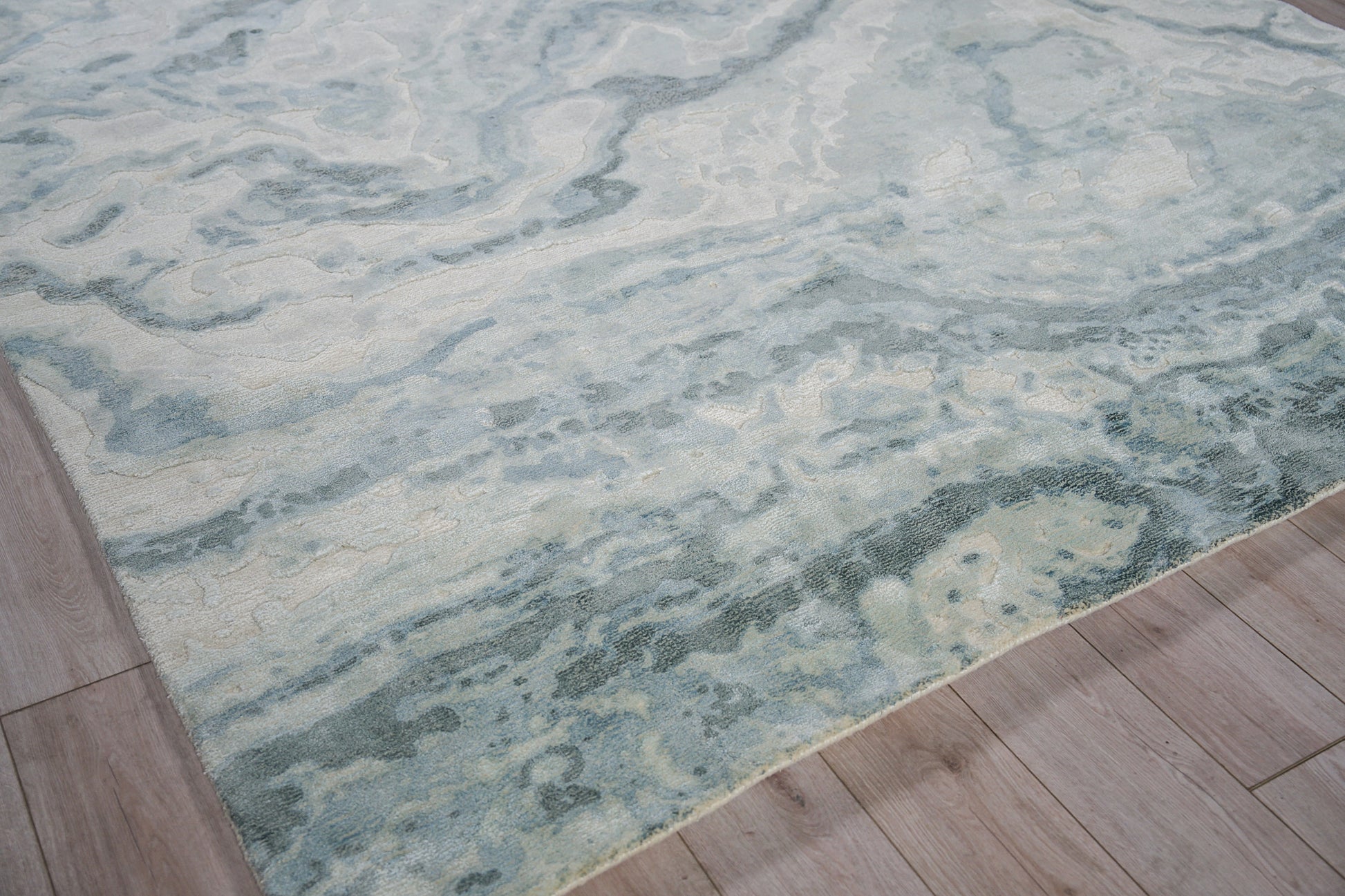 Rectangular blue-teal abstract area rug with ivory accents, water-like pattern, on light wood floor.