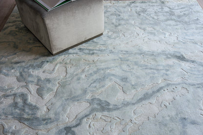 Light blue-gray abstract area rug with a plush pile under a gray fabric ottoman, with a magazine on top.