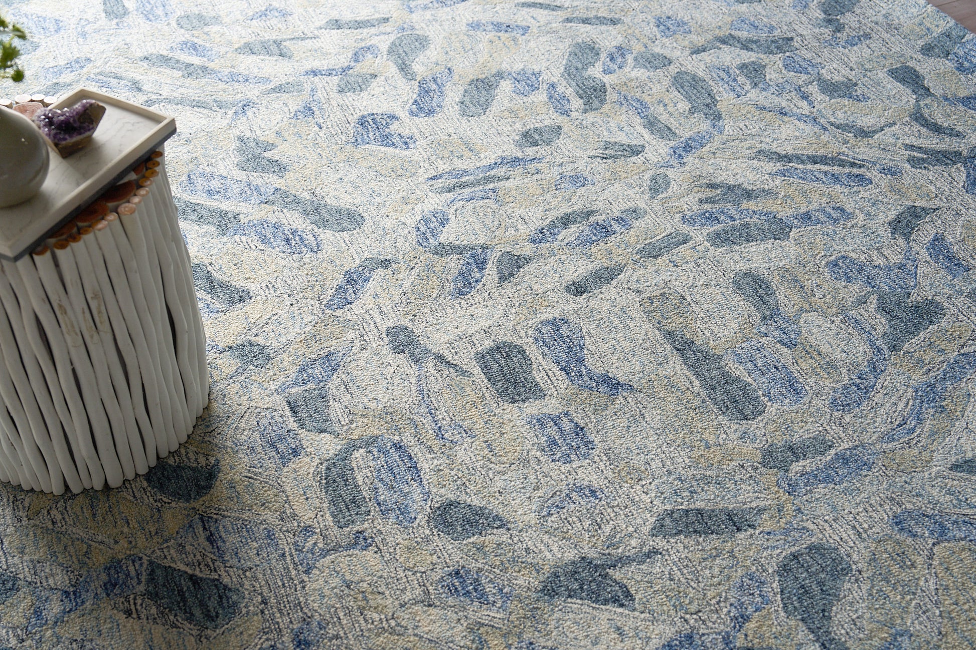Blue-gray stone-pattern rug with beige flecks; white woven side table on the left with decorative stones.