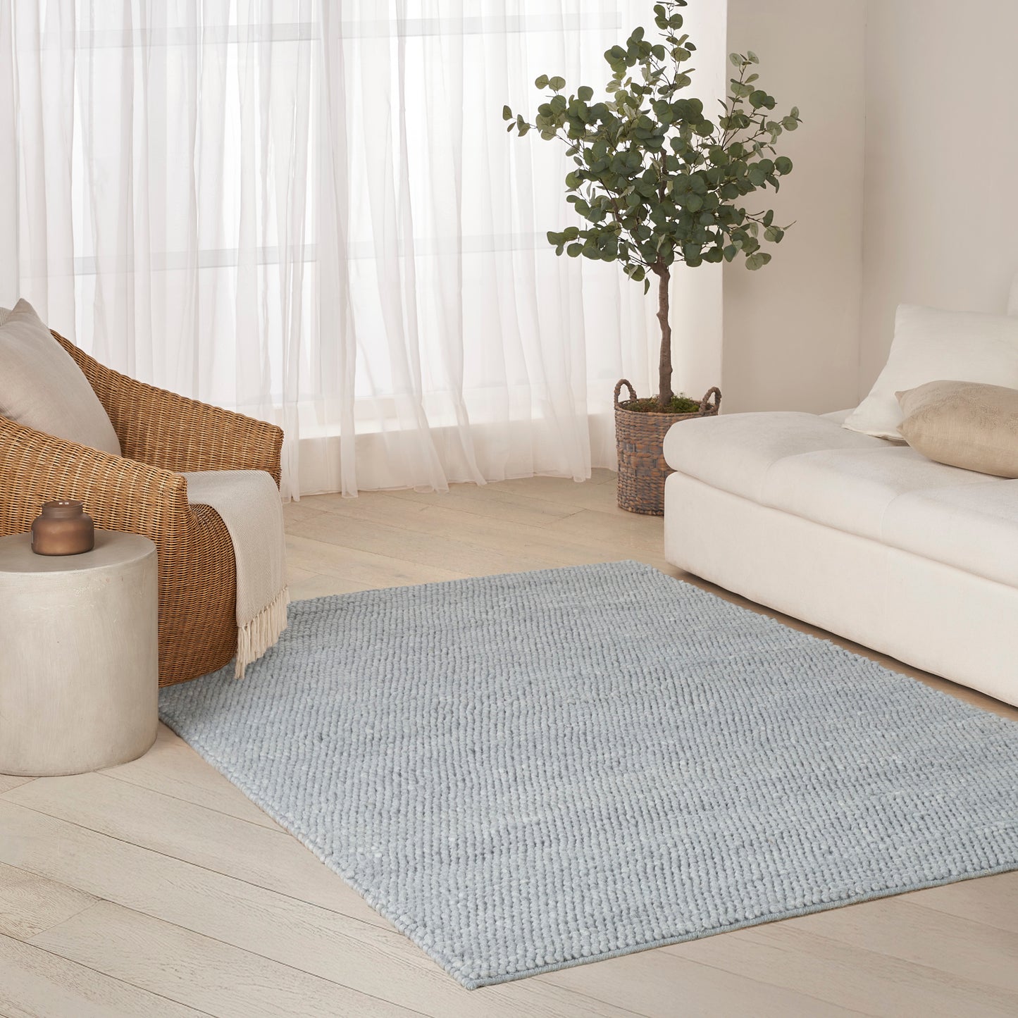Light blue textured rug on pale wood floor beside white sofa and wicker armchair with a potted plant and sheer curtains.