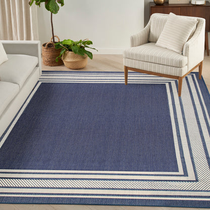 Navy blue woven area rug with light striped border, beige armchair with wooden legs, plant accents in a cozy living room.