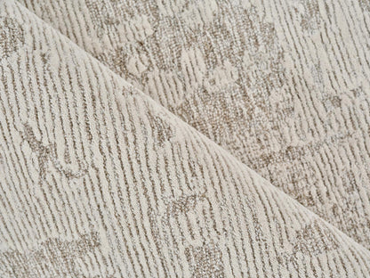 Beige textured rug close-up with subtle damask pattern and soft pile; folded edge visible.