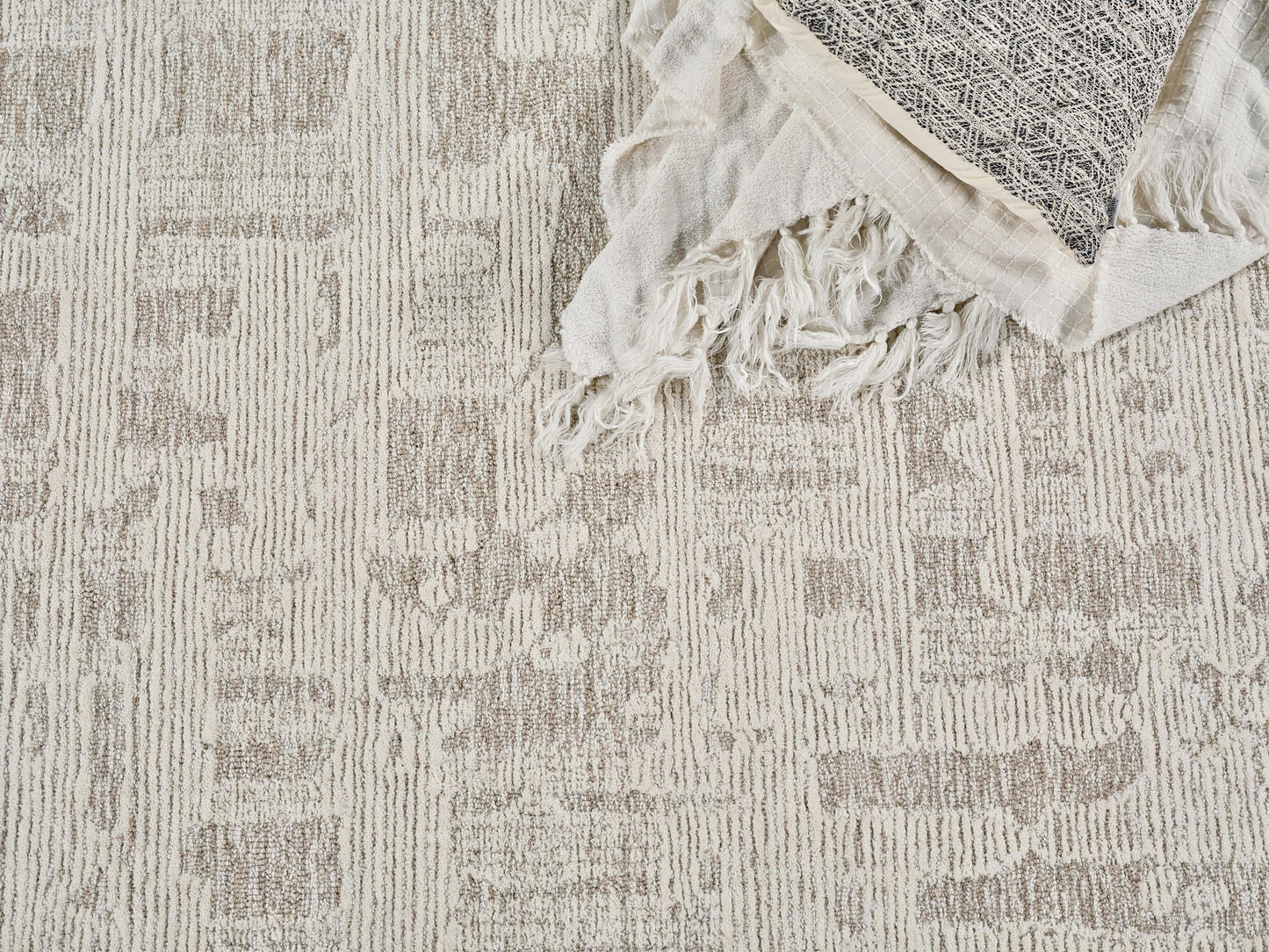 Neutral beige patterned rug with textured stripes, topped by a cream fringed throw with a dark geometric panel.