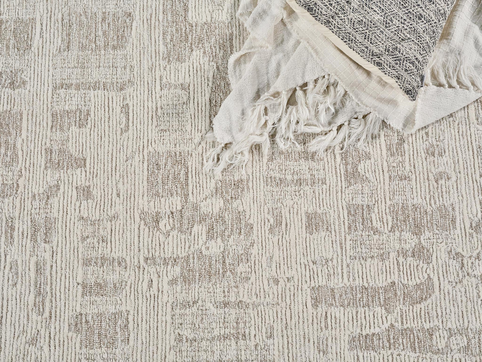 Neutral beige patterned rug with textured stripes, topped by a cream fringed throw with a dark geometric panel.