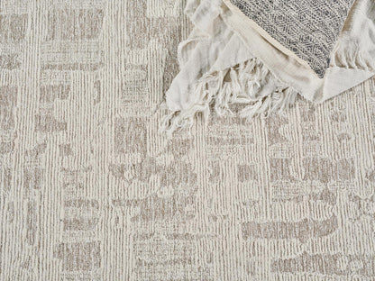 Neutral beige patterned rug with textured stripes, topped by a cream fringed throw with a dark geometric panel.
