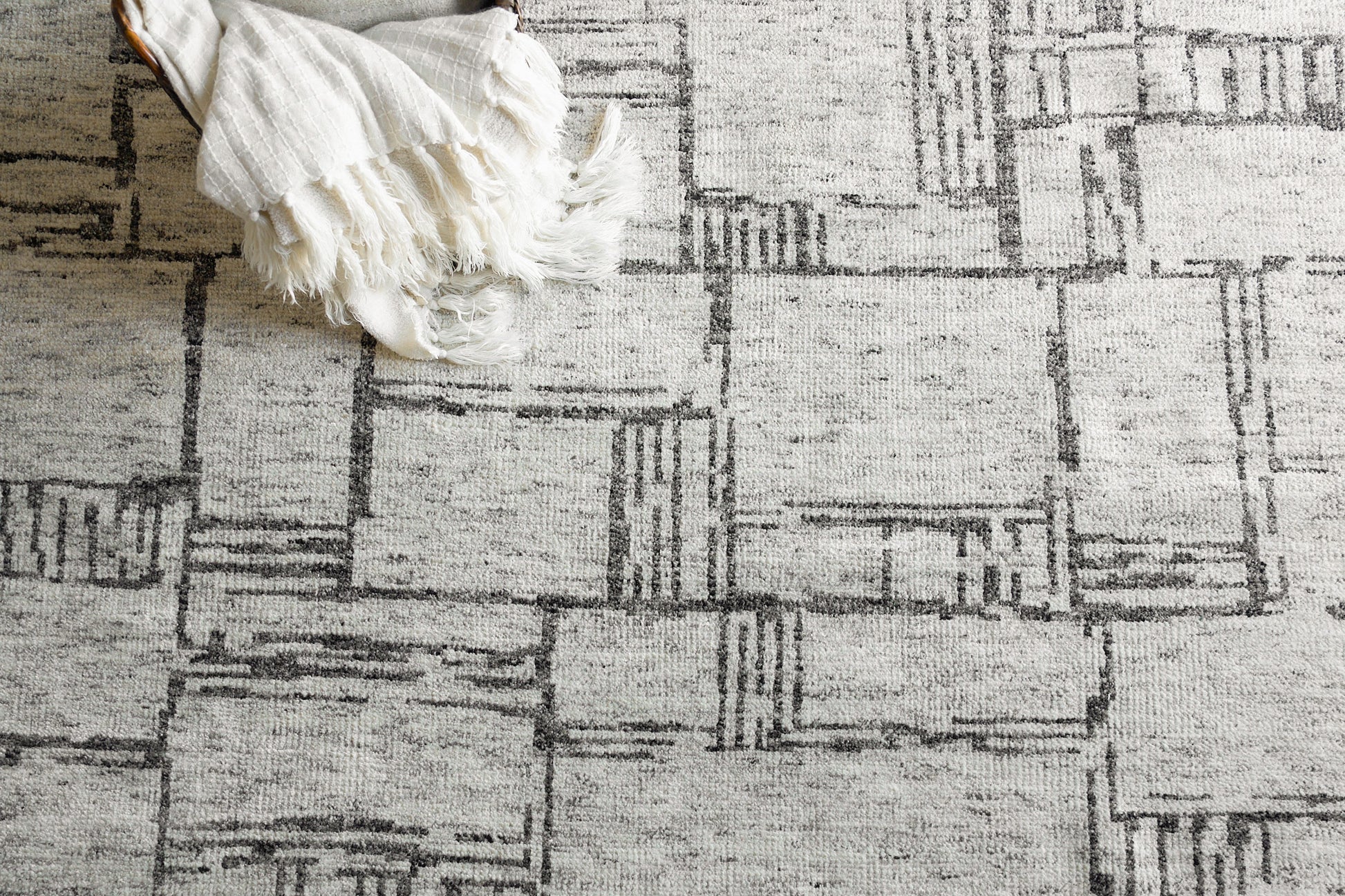 Cream and gray geometric patchwork hand-knotted rug with fringed edges, with a white blanket draped over a corner.