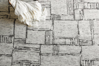 Cream and gray geometric patchwork hand-knotted rug with fringed edges, with a white blanket draped over a corner.