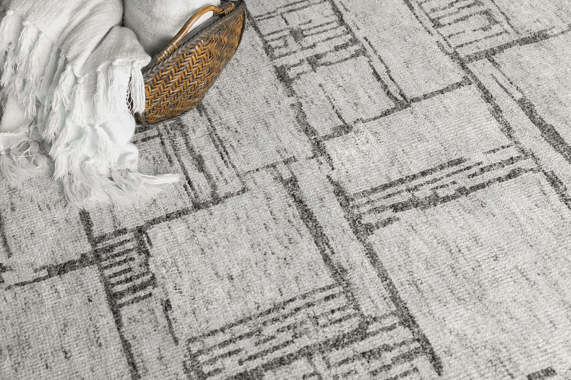 Grey abstract-pattern rug with geometric linear blocks; wicker basket and white fringed throw at edge.