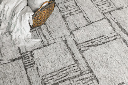 Grey abstract-pattern rug with geometric linear blocks; wicker basket and white fringed throw at edge.