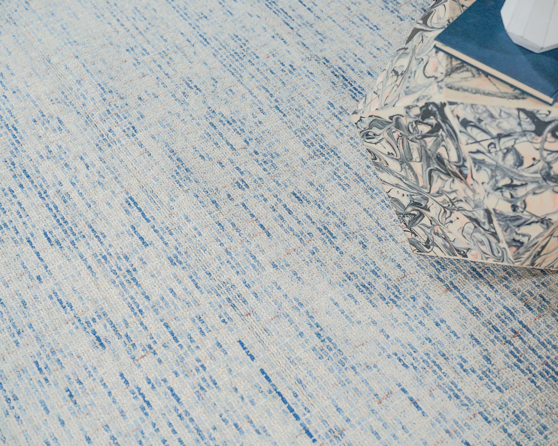 A light blue and ivory hand-tufted rug with subtle blue streaks, beside a marble-patterned cube ottoman and a blue notebook.
